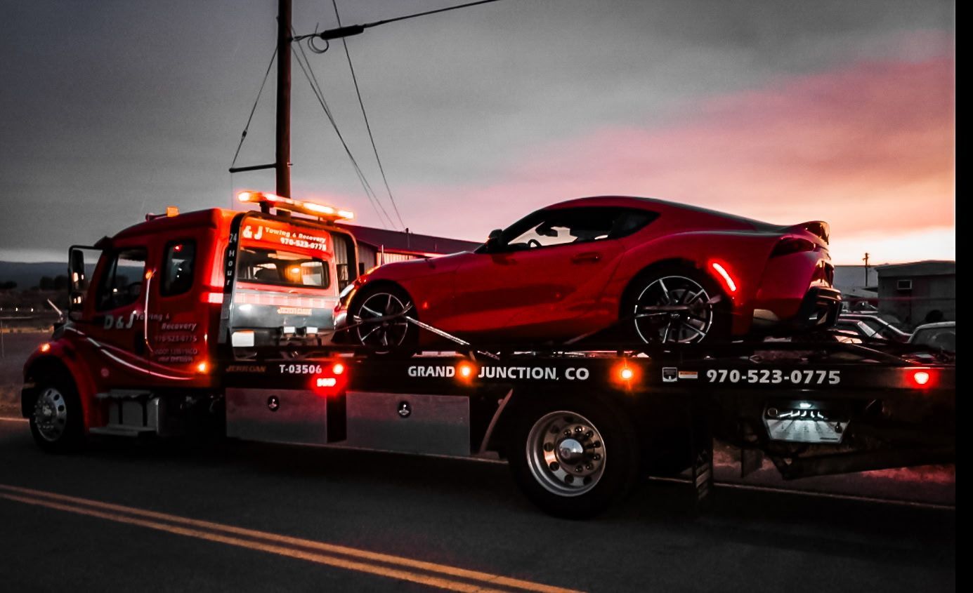 Red sports car on a tow truck at dusk. Truck has flashing lights.
