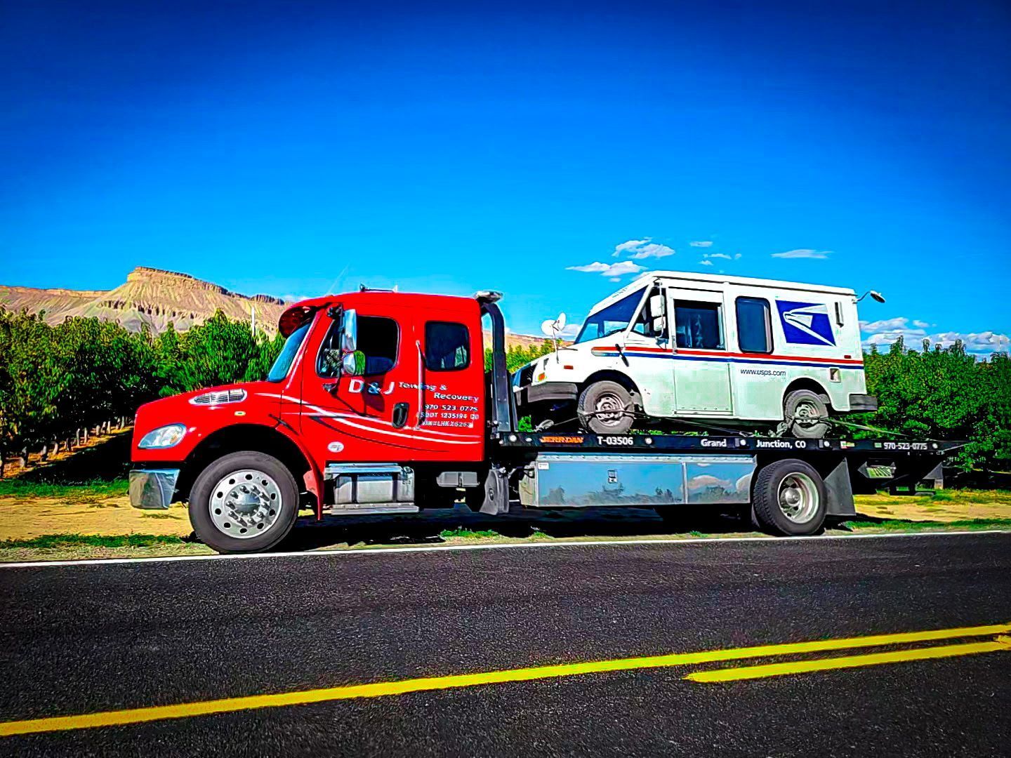 Red tow truck hauling a US Postal Service mail truck on a sunny road.