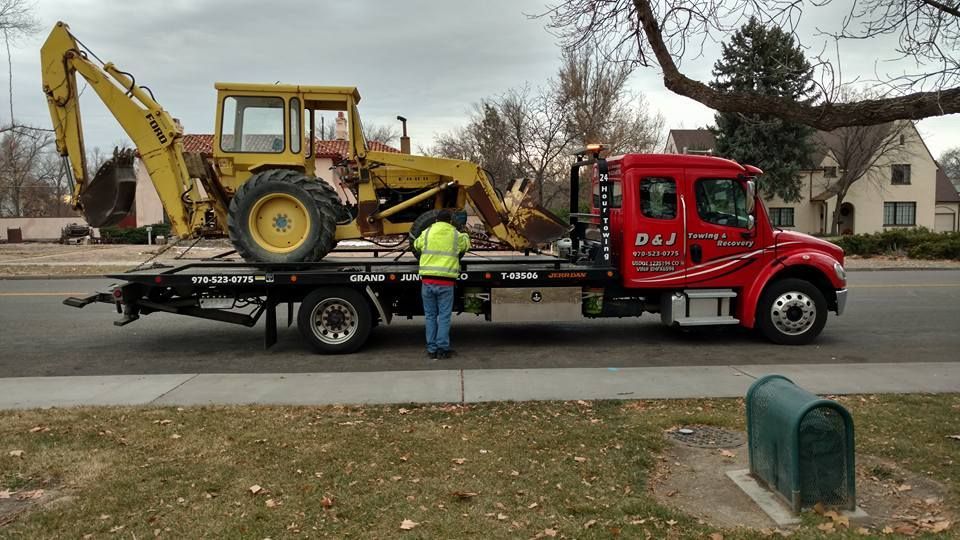 Yellow backhoe loaded on a tow truck by a person in a safety vest, roadside scene.