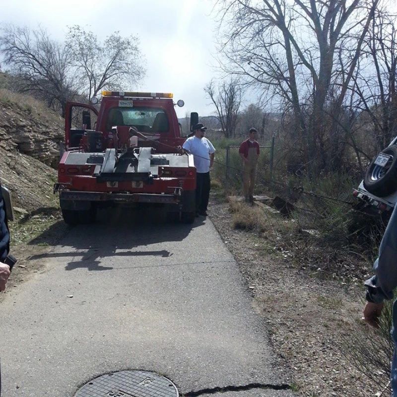 Tow truck on narrow path, vehicle being recovered. Two men watch, trees in background.