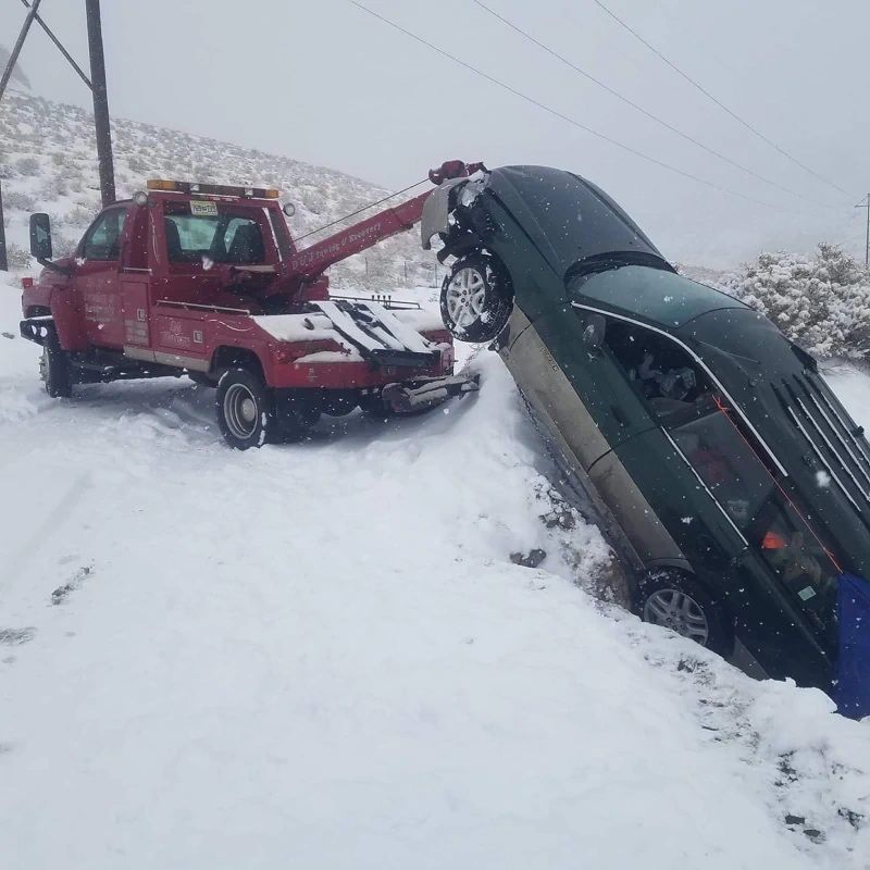 Red tow truck pulling a green car out of a snow-filled ditch on a snowy roadside.