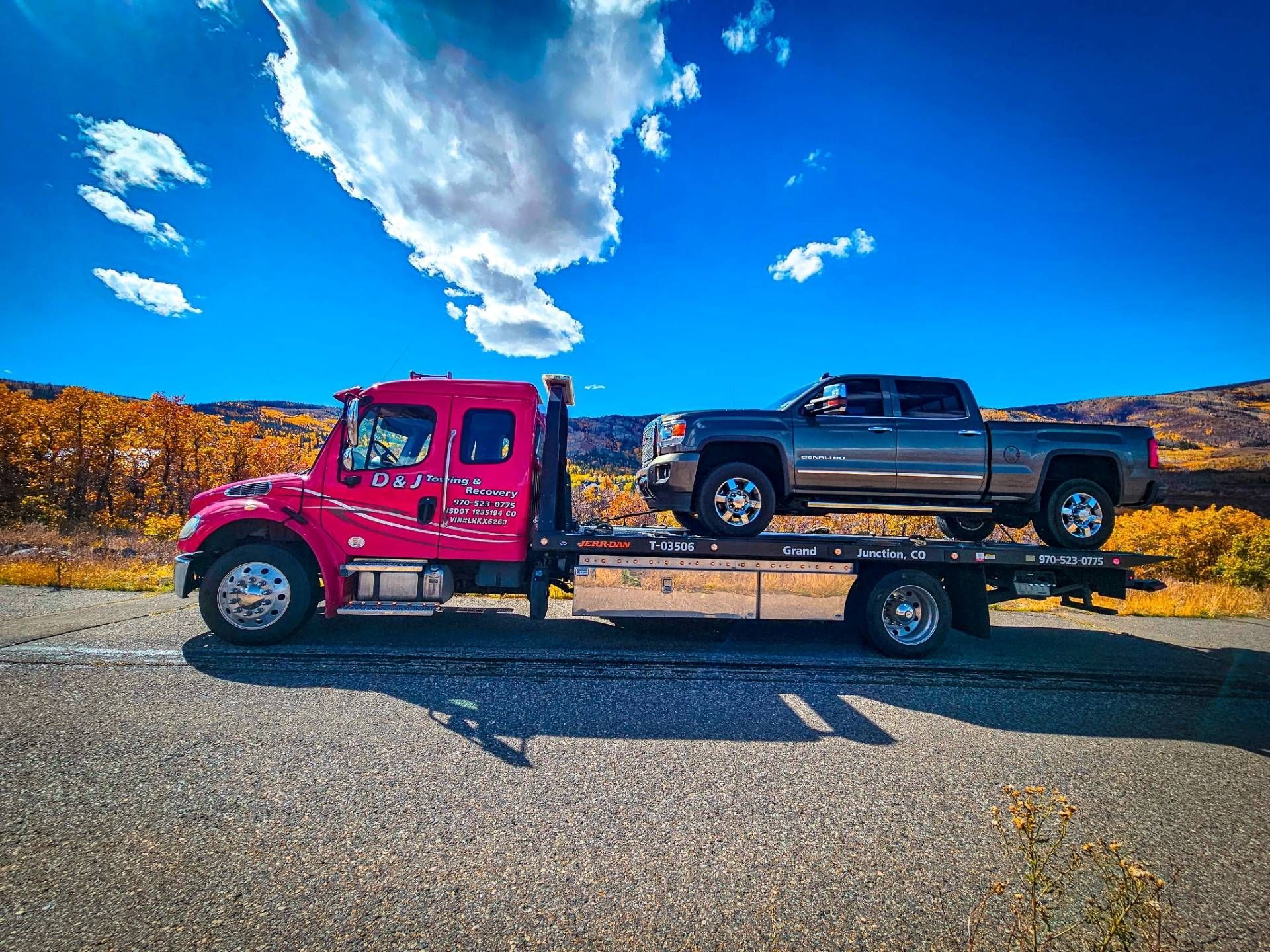 Pink tow truck carrying a gray pickup truck on a road. Autumn trees under a blue sky with clouds.