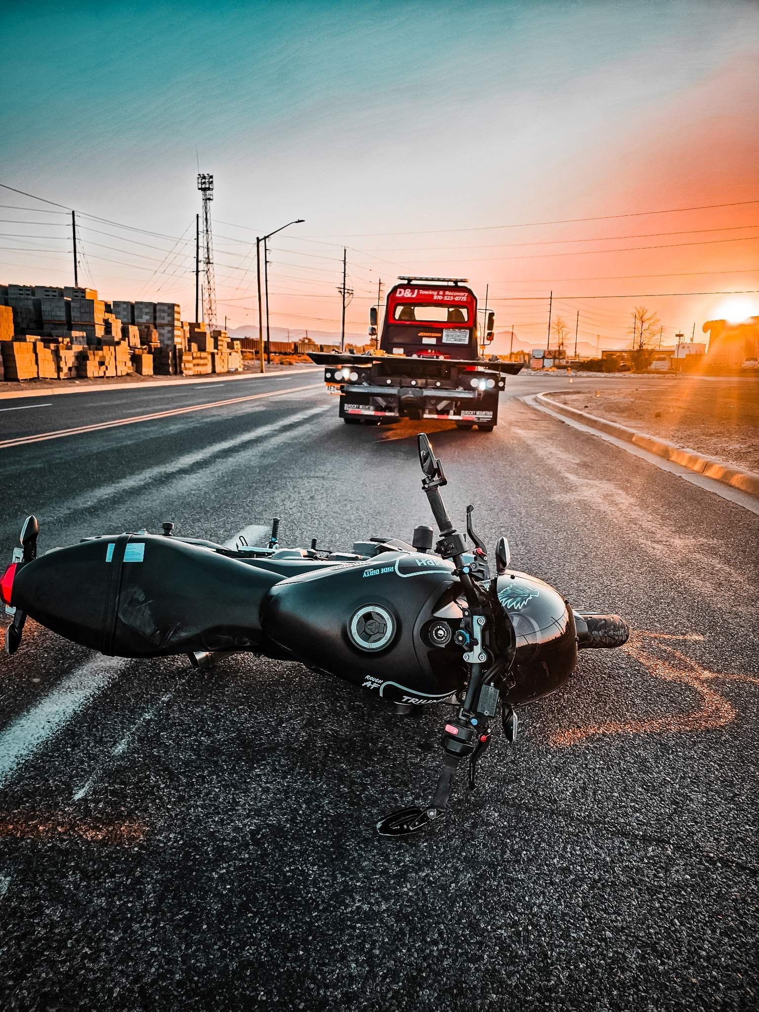 Motorcycle on pavement after an accident, tow truck approaching. Sunset in the background.