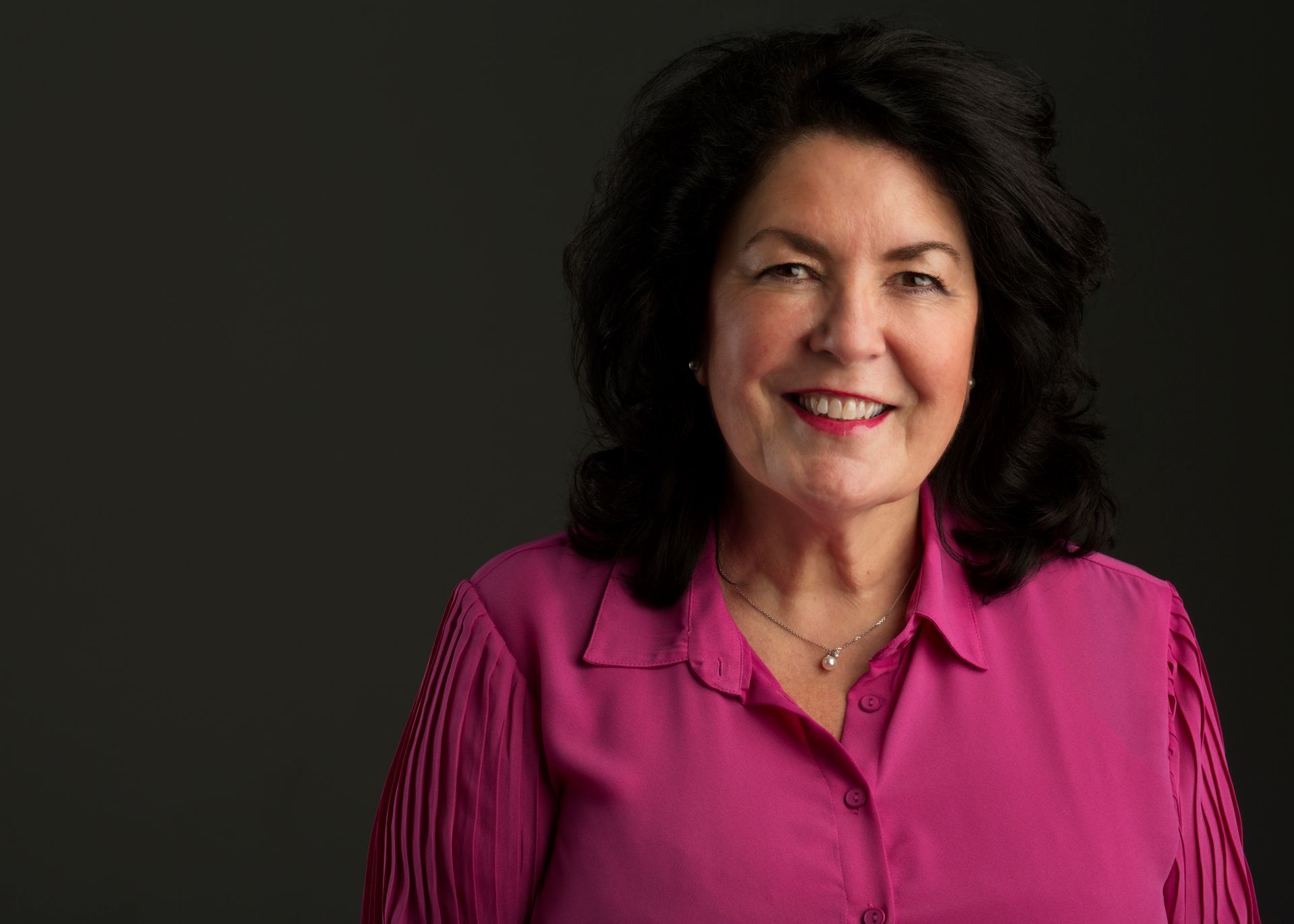 A person with dark, wavy hair smiling, wearing a vibrant pink collared shirt and a simple necklace against a dark background.