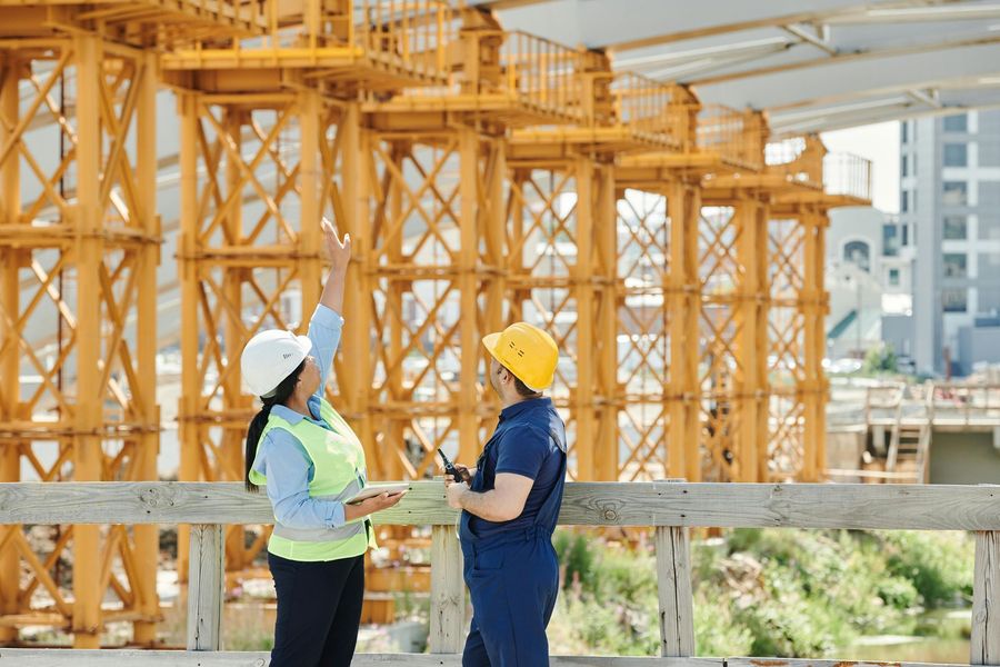 Two construction workers in safety gear stand on a bridge, looking up and discussing the yellow scaffolding structure.