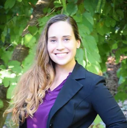 Alicia in a black jacket and purple shirt is smiling for the camera.