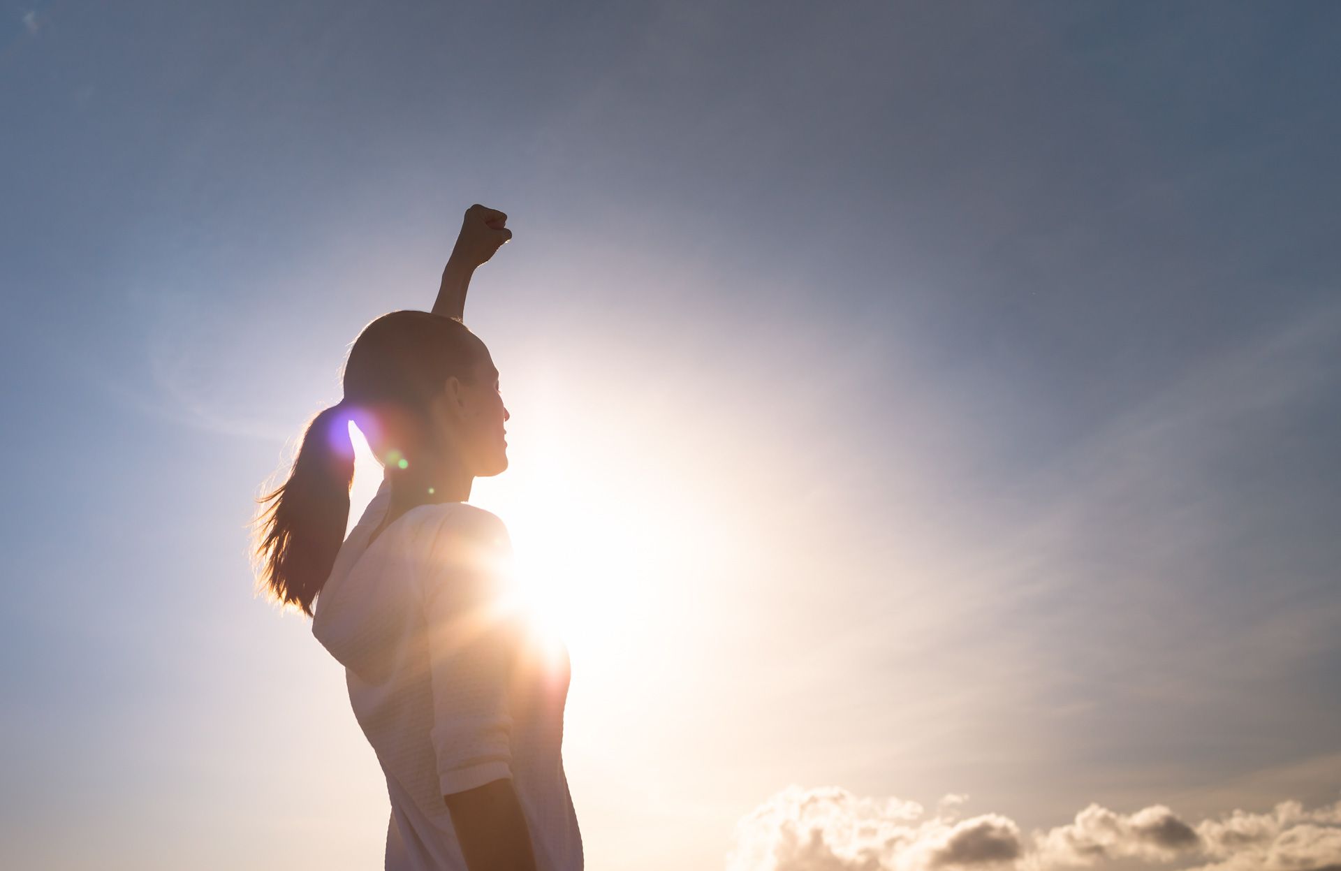 A woman is standing in front of the sun with her arms in the air.