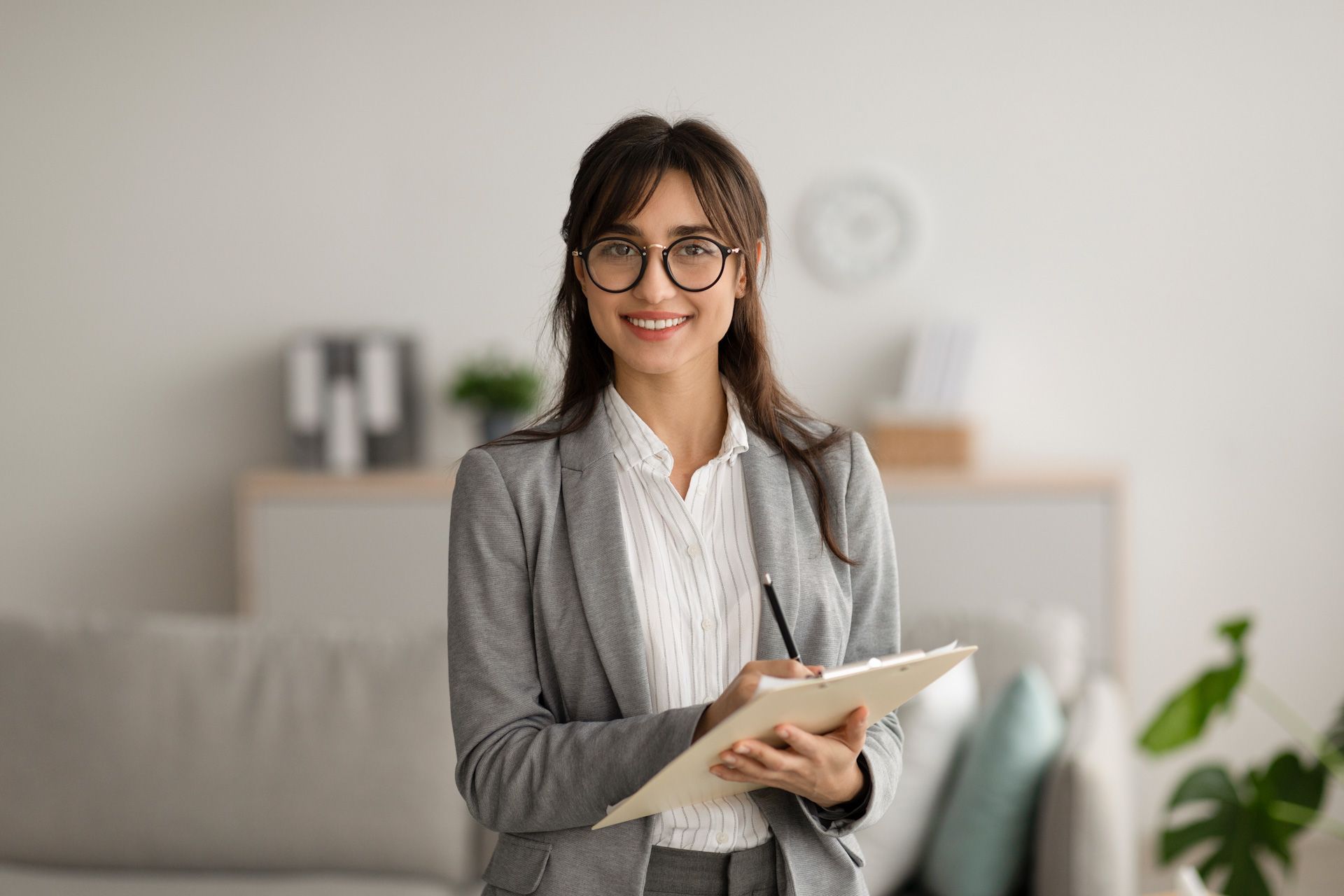 A woman in a suit and glasses is holding a clipboard and smiling.