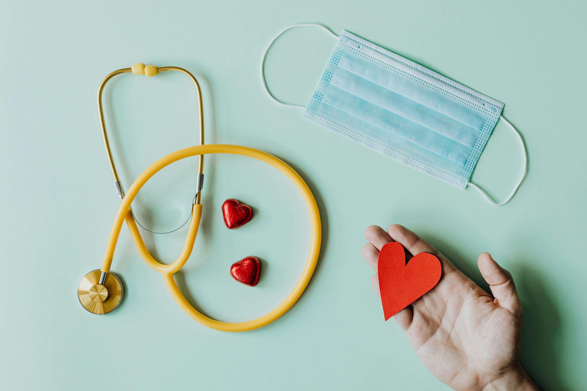 Yellow stethoscope, face mask, red hearts, and hand holding a red paper heart on a green surface.