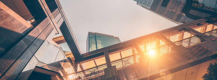 Low-angle view of modern city skyscrapers framing an elevated walkway illuminated by bright, warm sunlight.