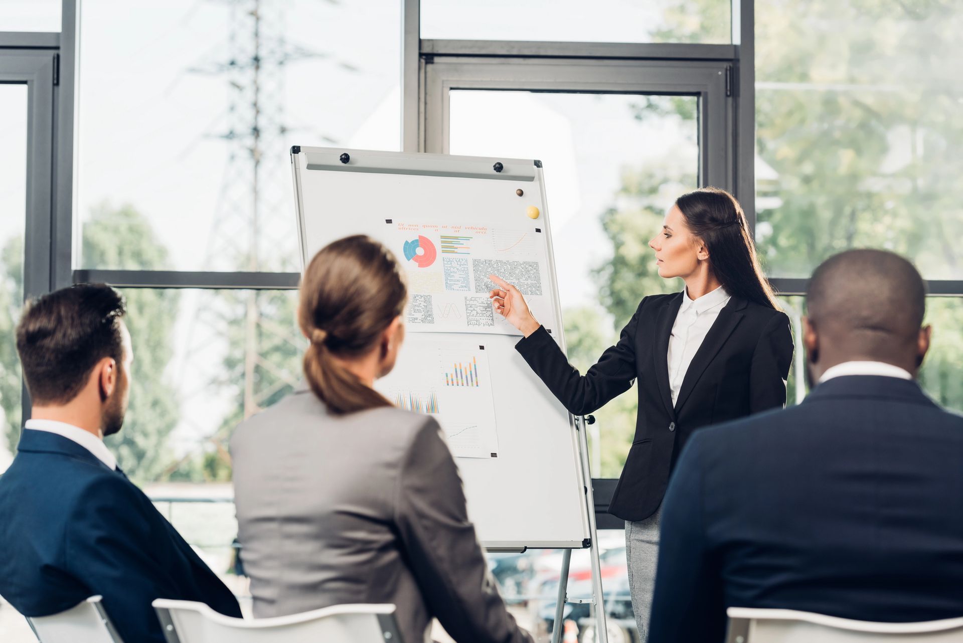 Una mujer con traje presenta un gráfico en una pizarra a un grupo pequeño en una oficina luminosa.