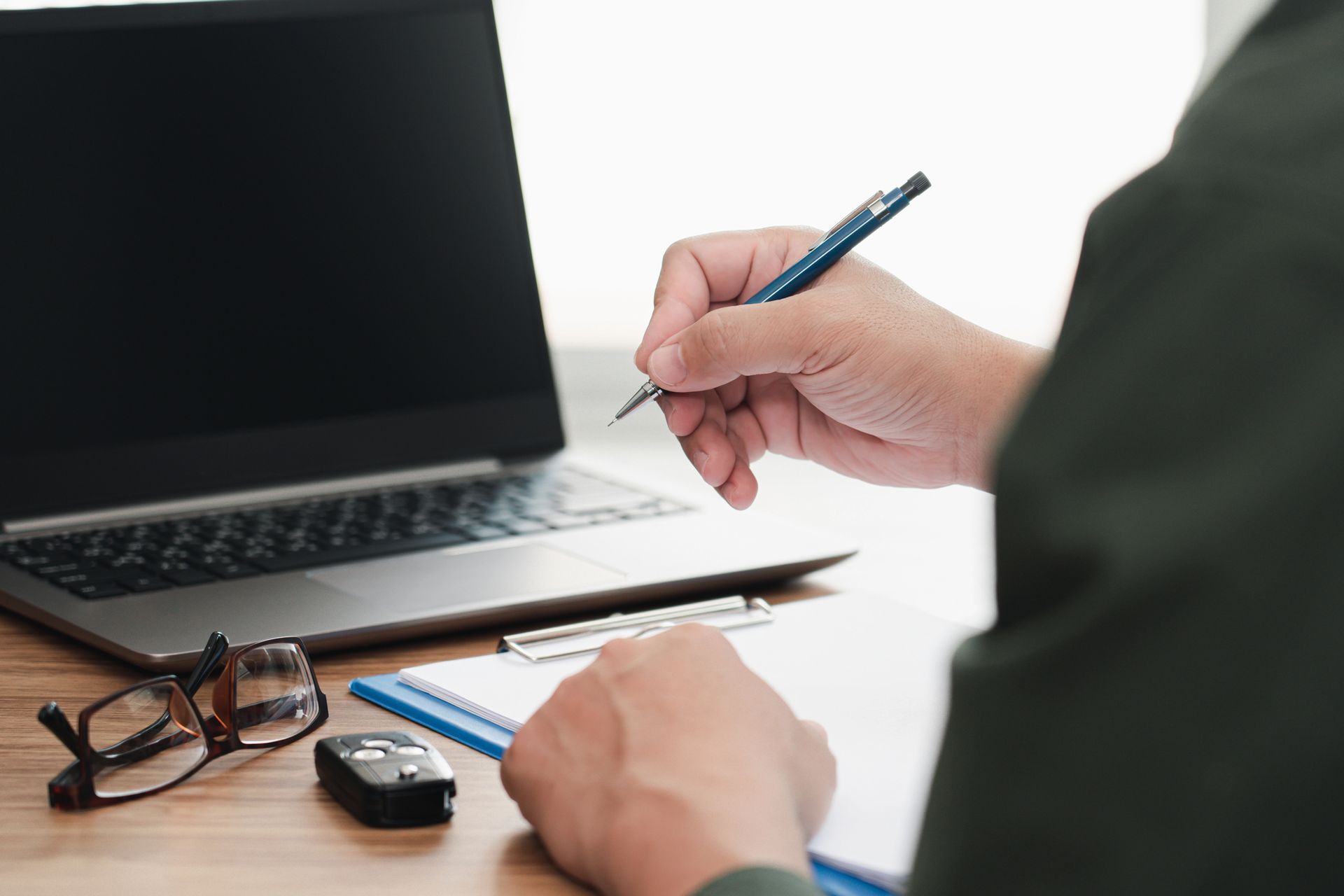 La persona escribe con un bolígrafo en un portapapeles, tiene una computadora portátil abierta