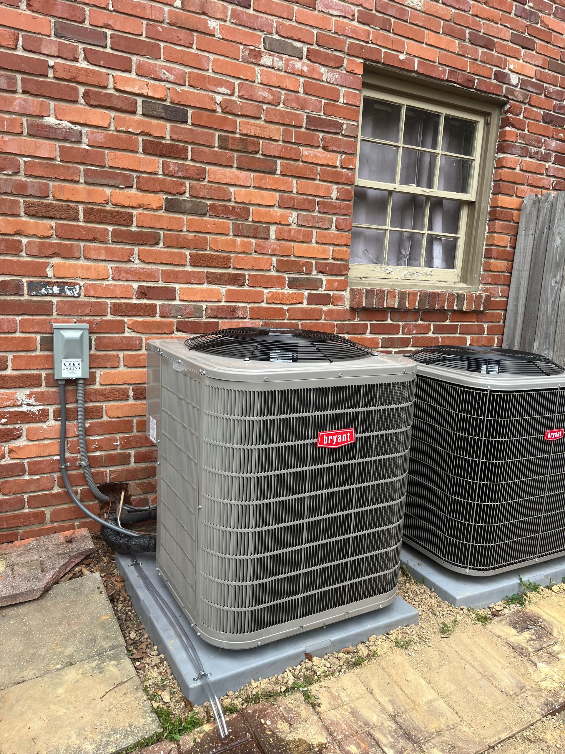 Two air conditioning units against a brick wall, near a window, with a power box.