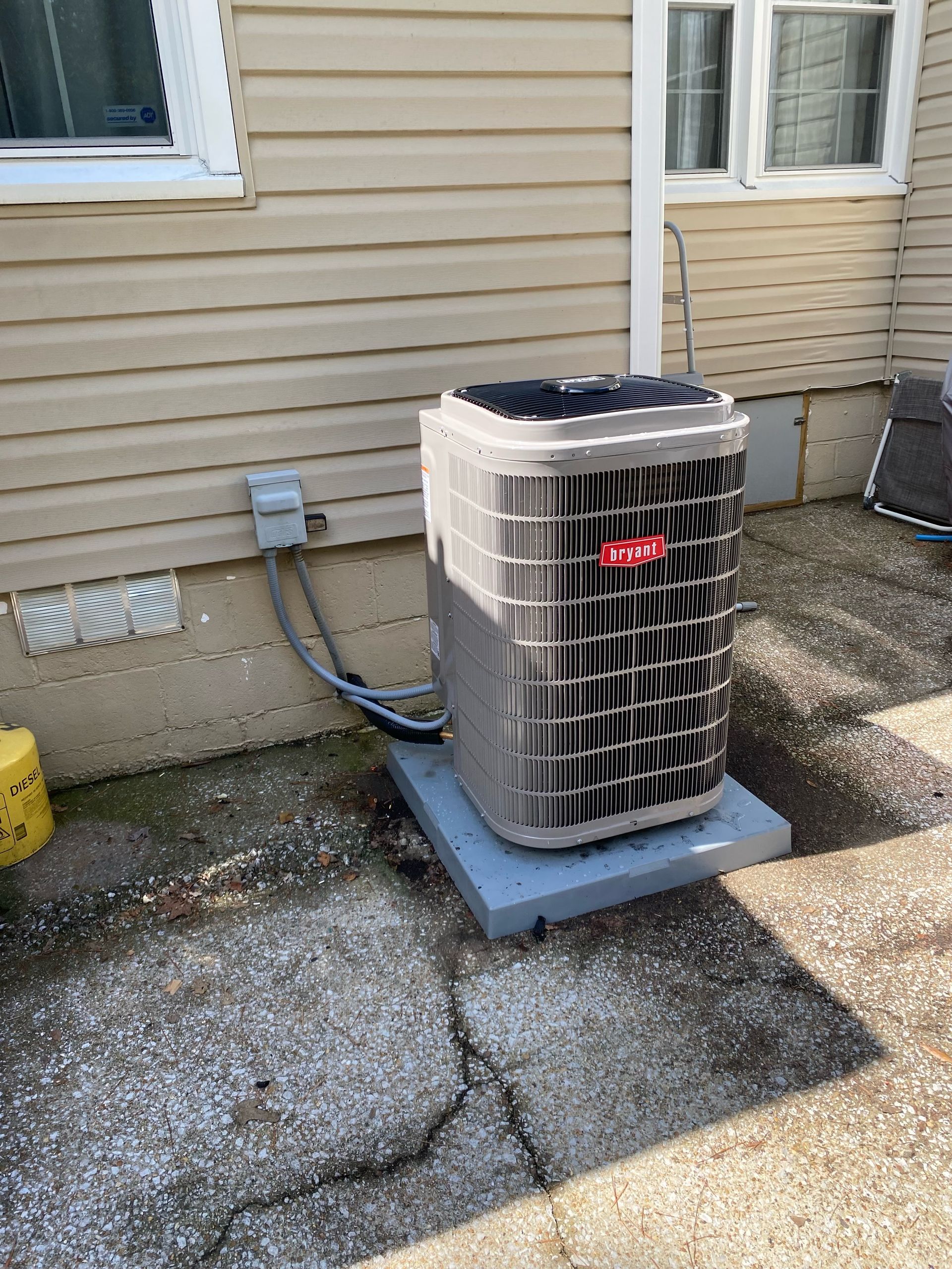 An outdoor Bryant air conditioning unit on a concrete pad next to a house with beige siding.