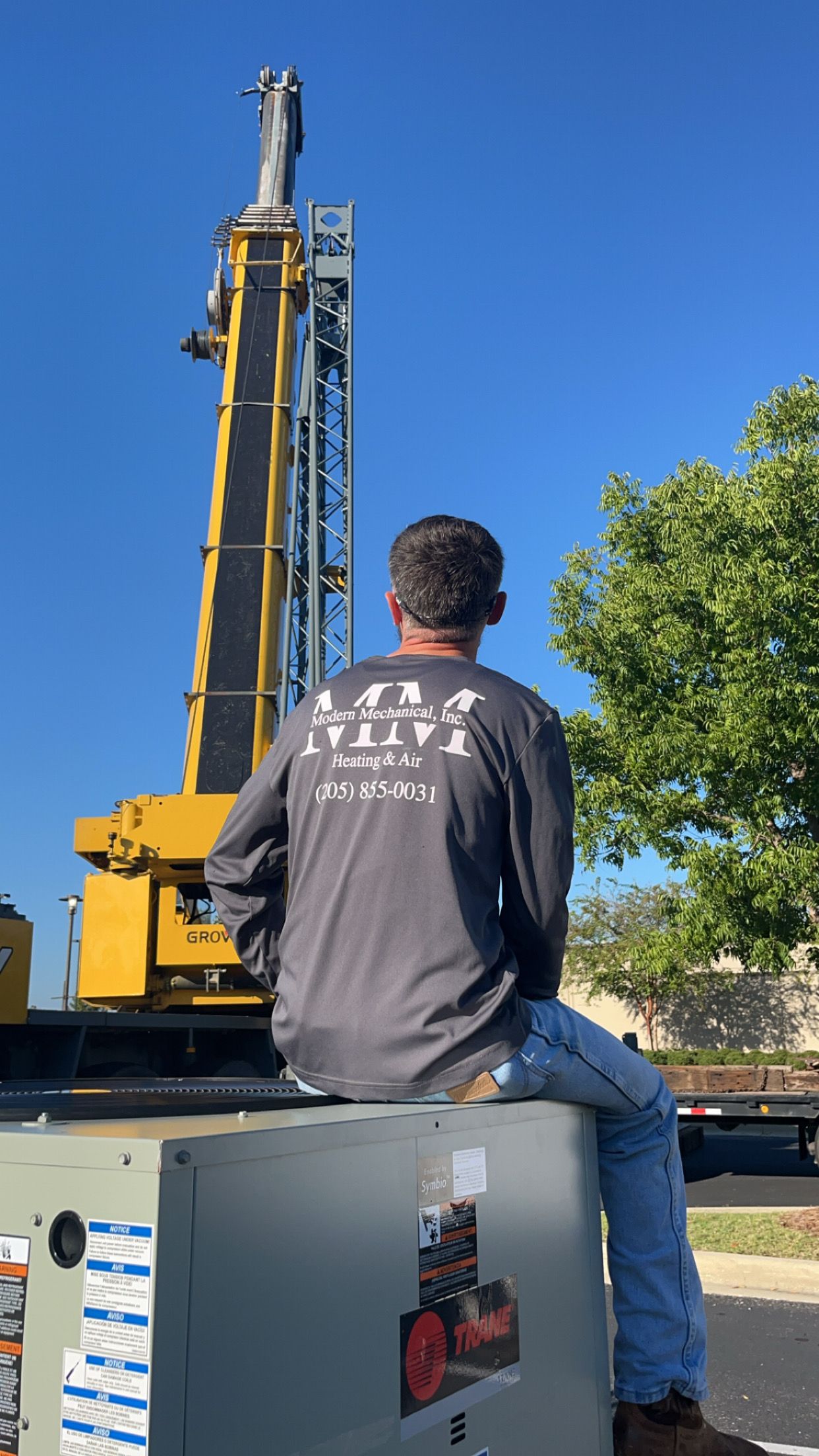 Man sits on a unit, looking up at a crane. Construction site with a blue sky and a tree.