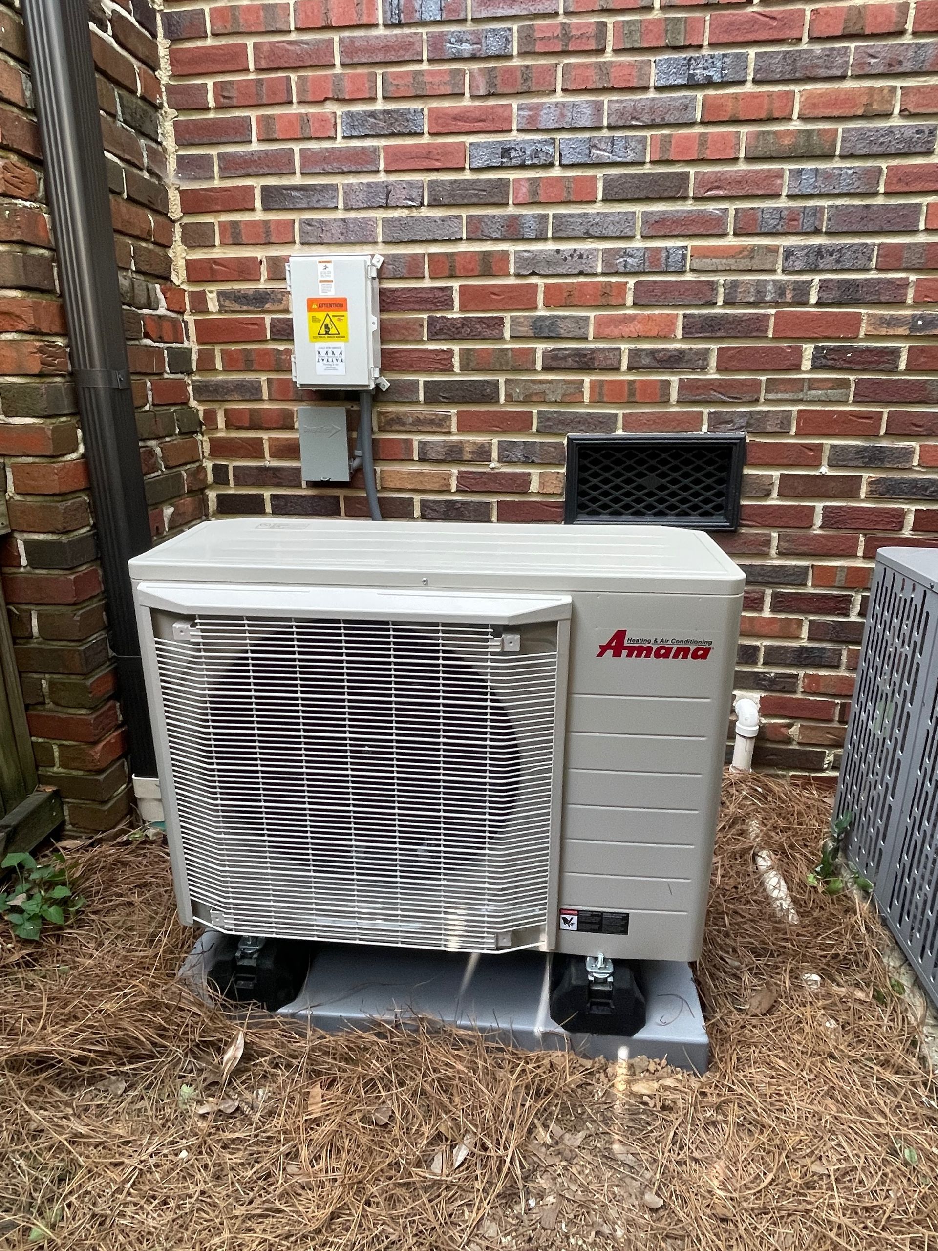 Air conditioner unit against a brick wall. Gray unit sits on a concrete pad with a warning sign above.