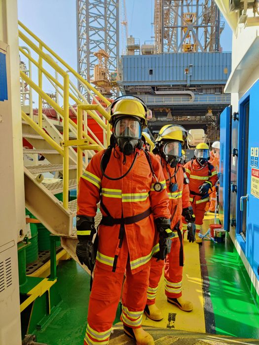 Four firefighters in orange protective suits and gas masks stand on a ship deck near a structure.
