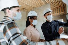 Three masked construction workers in hard hats examining plans at a construction site.