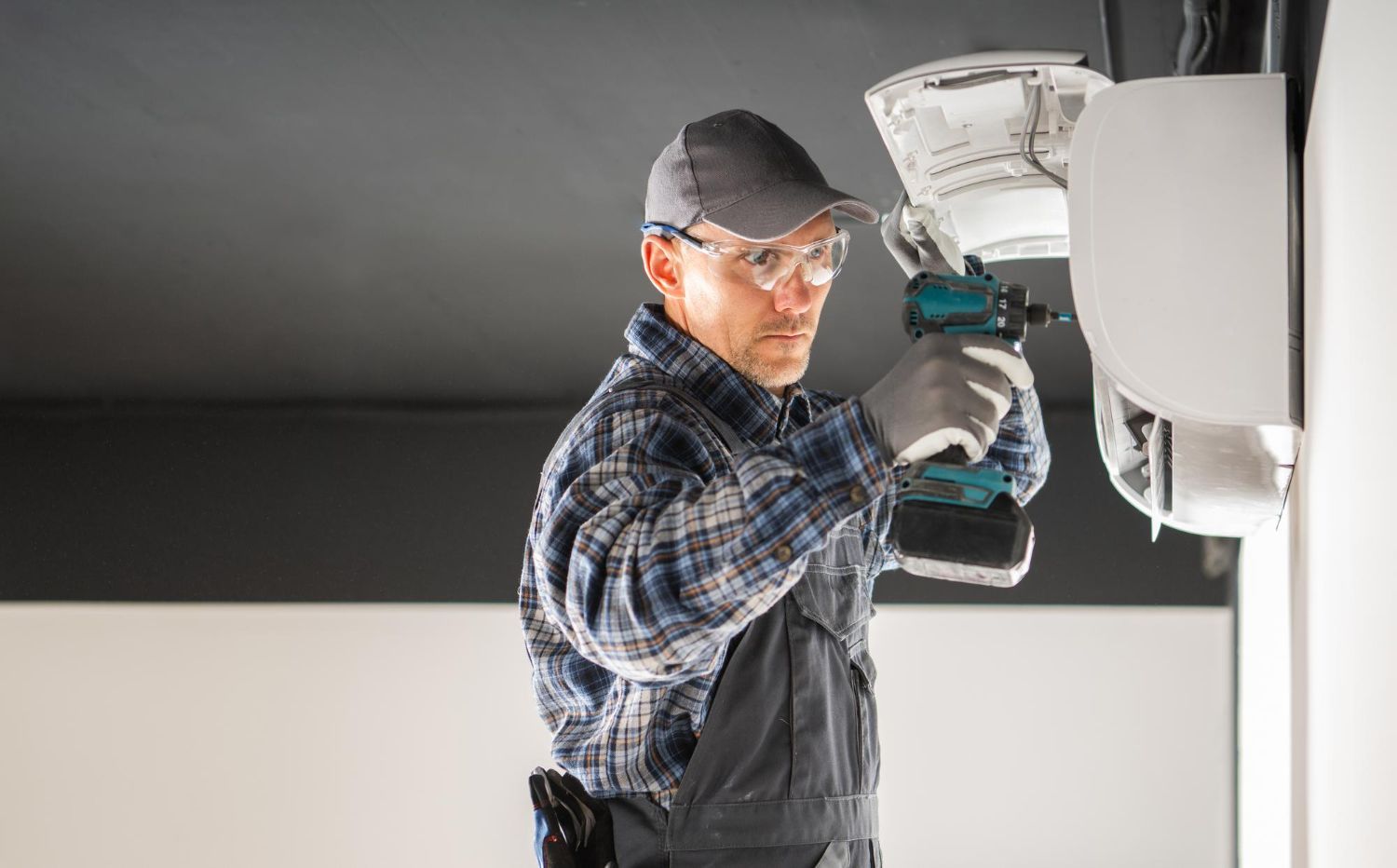 Electrician in a grey hat and safety glasses, drilling into an appliance on a white wall.