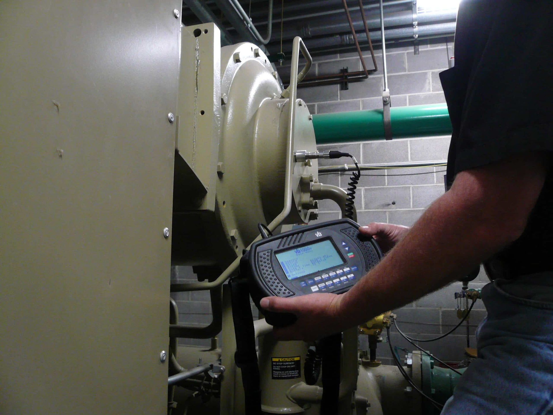 Person holding a diagnostic device, inspecting a large industrial machine in a mechanical room.