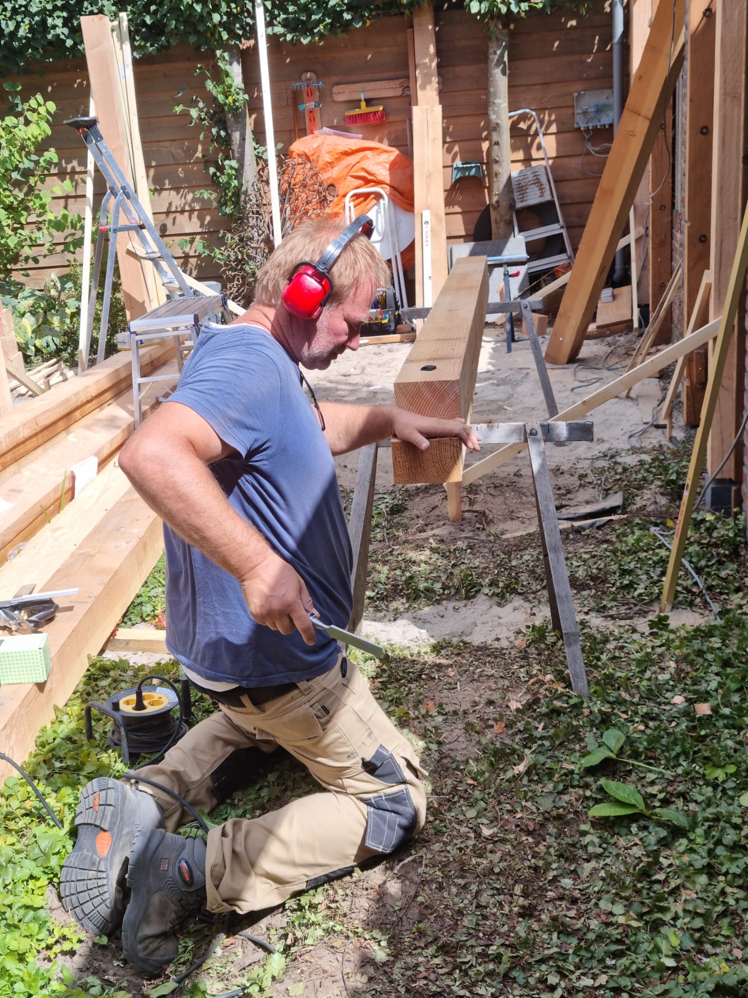 Een man met oorkappen zit op zijn knieën voor een stapel hout.