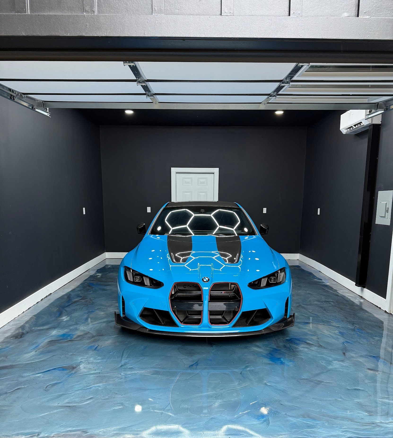 Blue BMW sports car in a black-walled garage with a blue, epoxy-like floor, lit by overhead lights.
