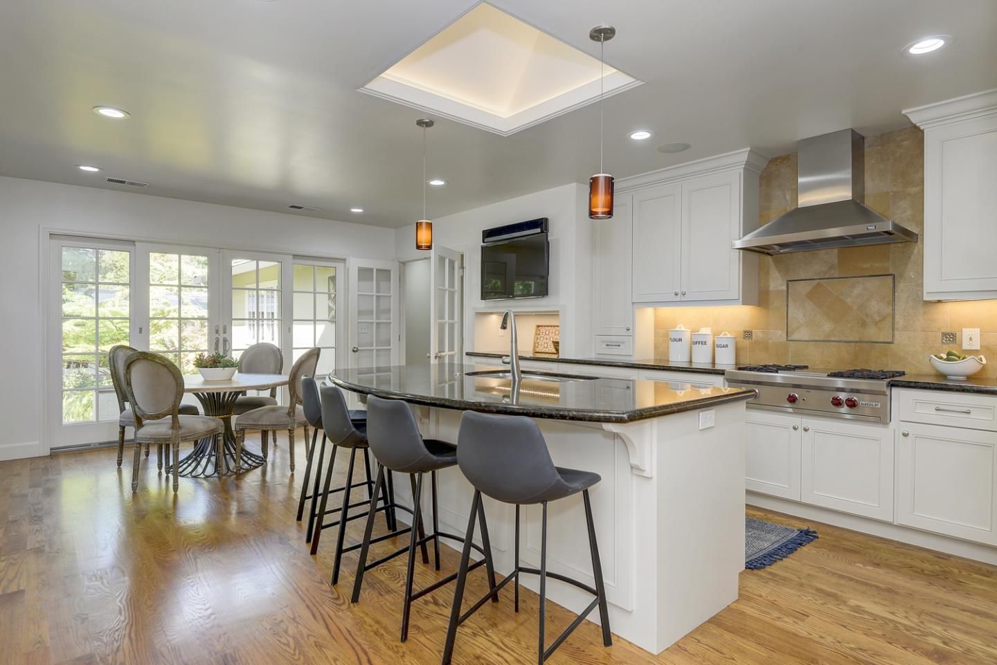 Spacious white kitchen with island, stools, stainless steel appliances, and dining area with table and chairs.