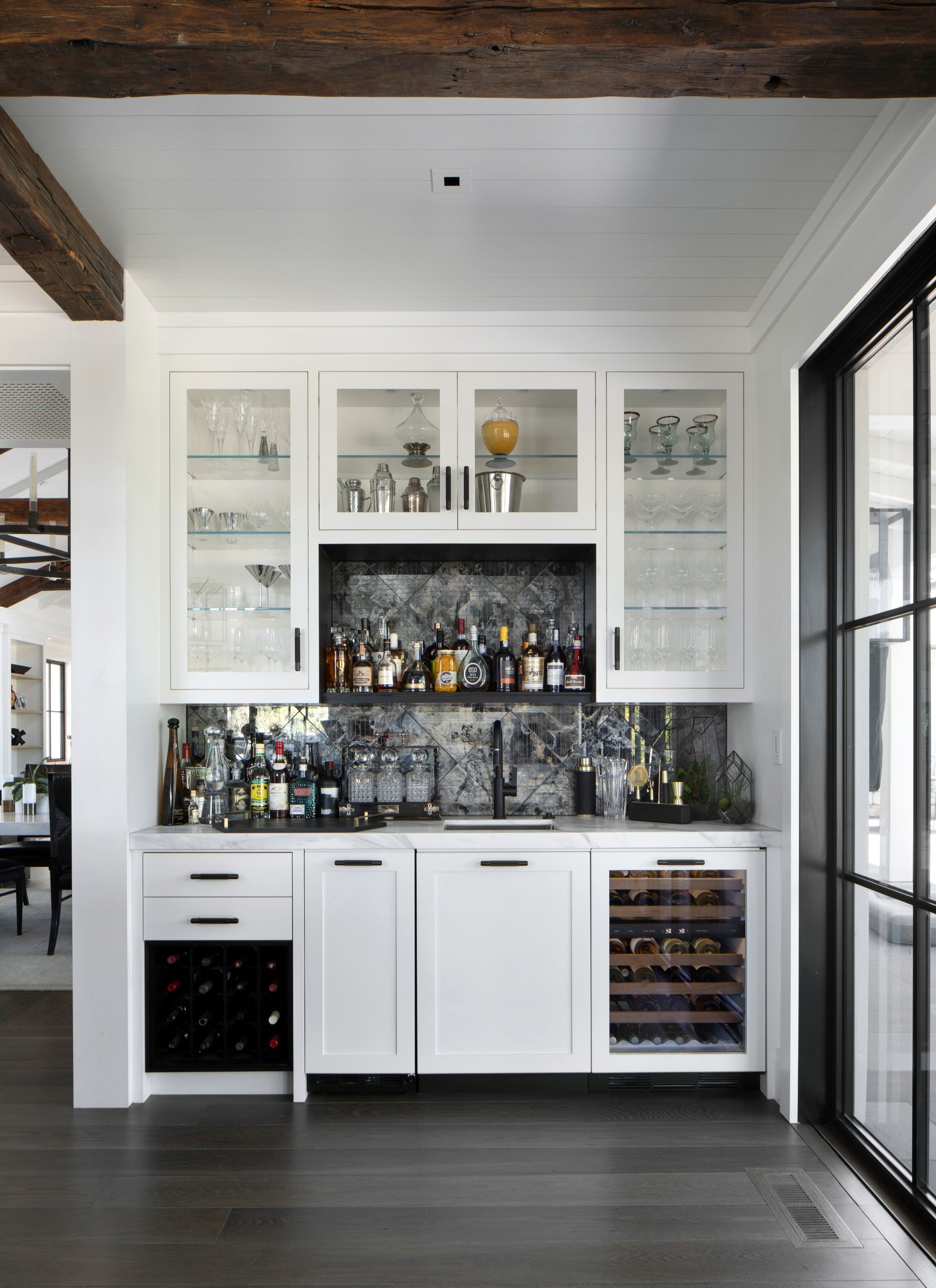 White built-in bar with mirrored backsplash, liquor, glassware, and wine coolers. Set in a modern, bright room.