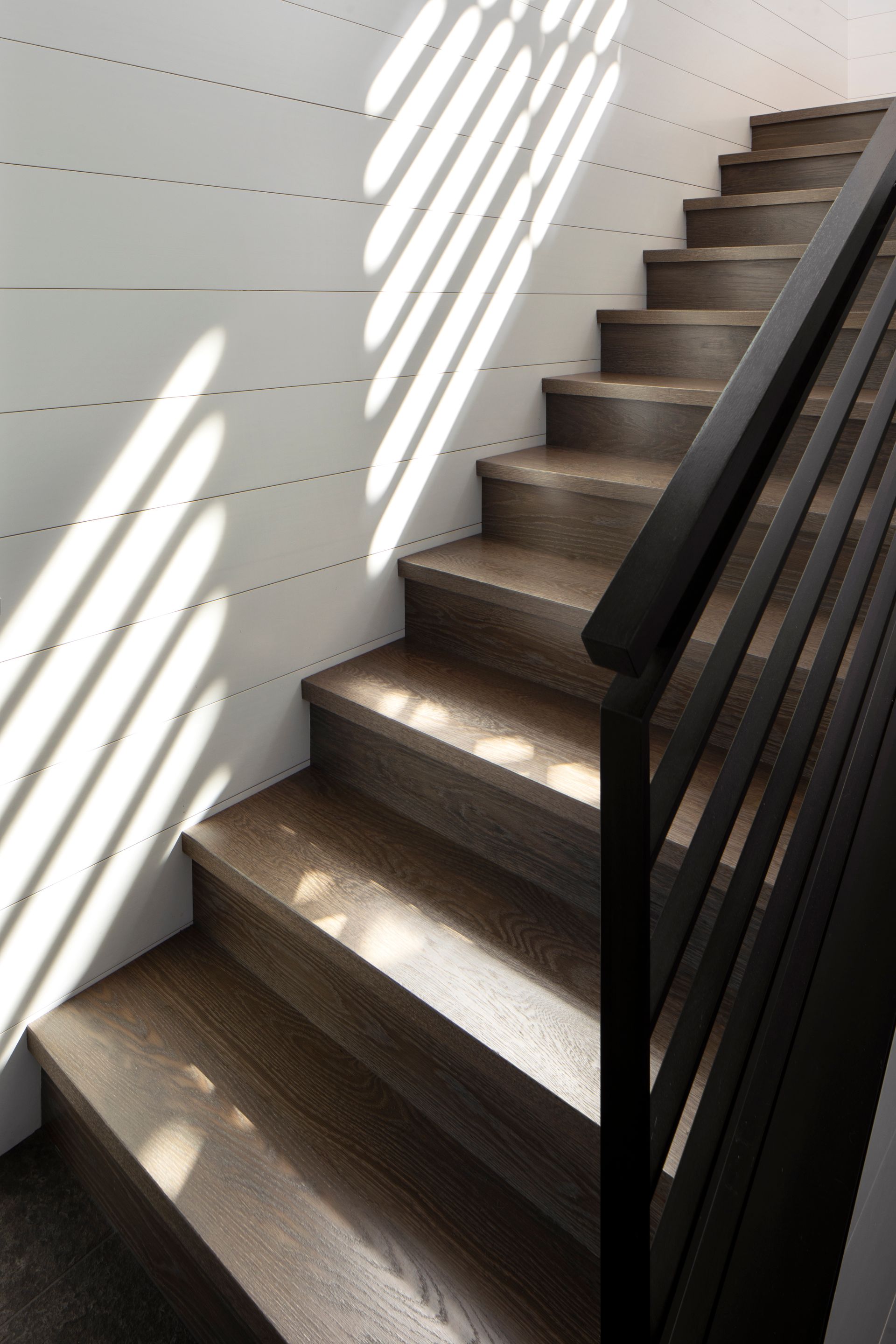 Wooden staircase with dark handrail, sunlight and shadows on white wall.