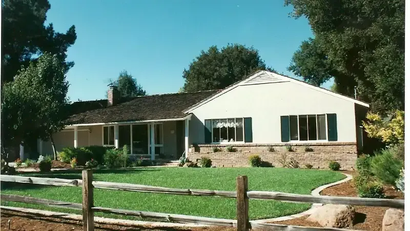 Ranch-style house with beige siding, brown roof, green lawn, wooden fence, and blue sky.