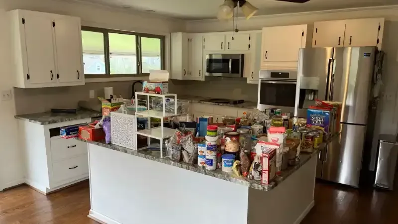 A cluttered kitchen with white cabinets, island, and stainless steel fridge overflowing with groceries.