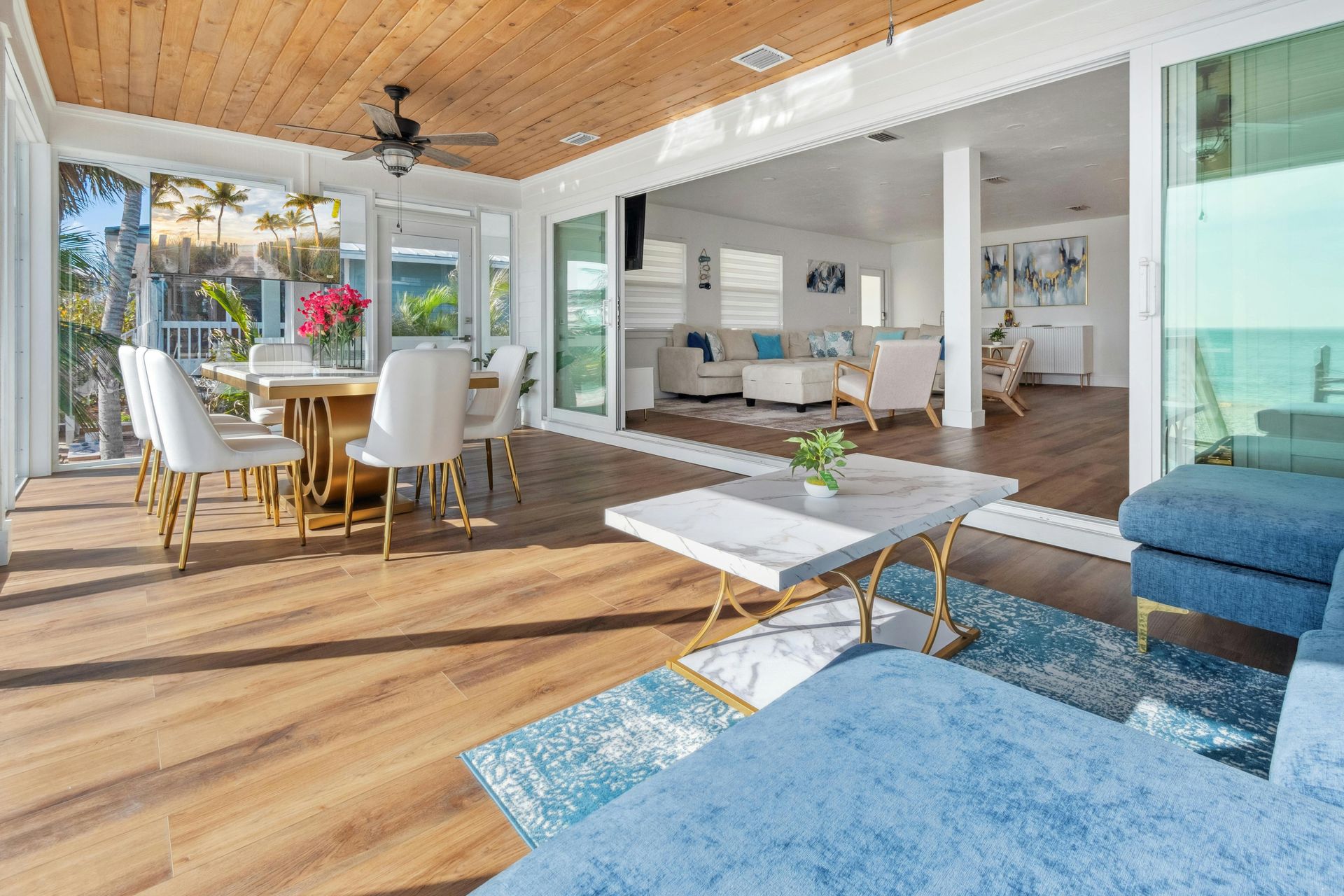 An open-concept room with wood ceilings, a dining area, and a blue sofa, leading to a bright, windowed sunroom.