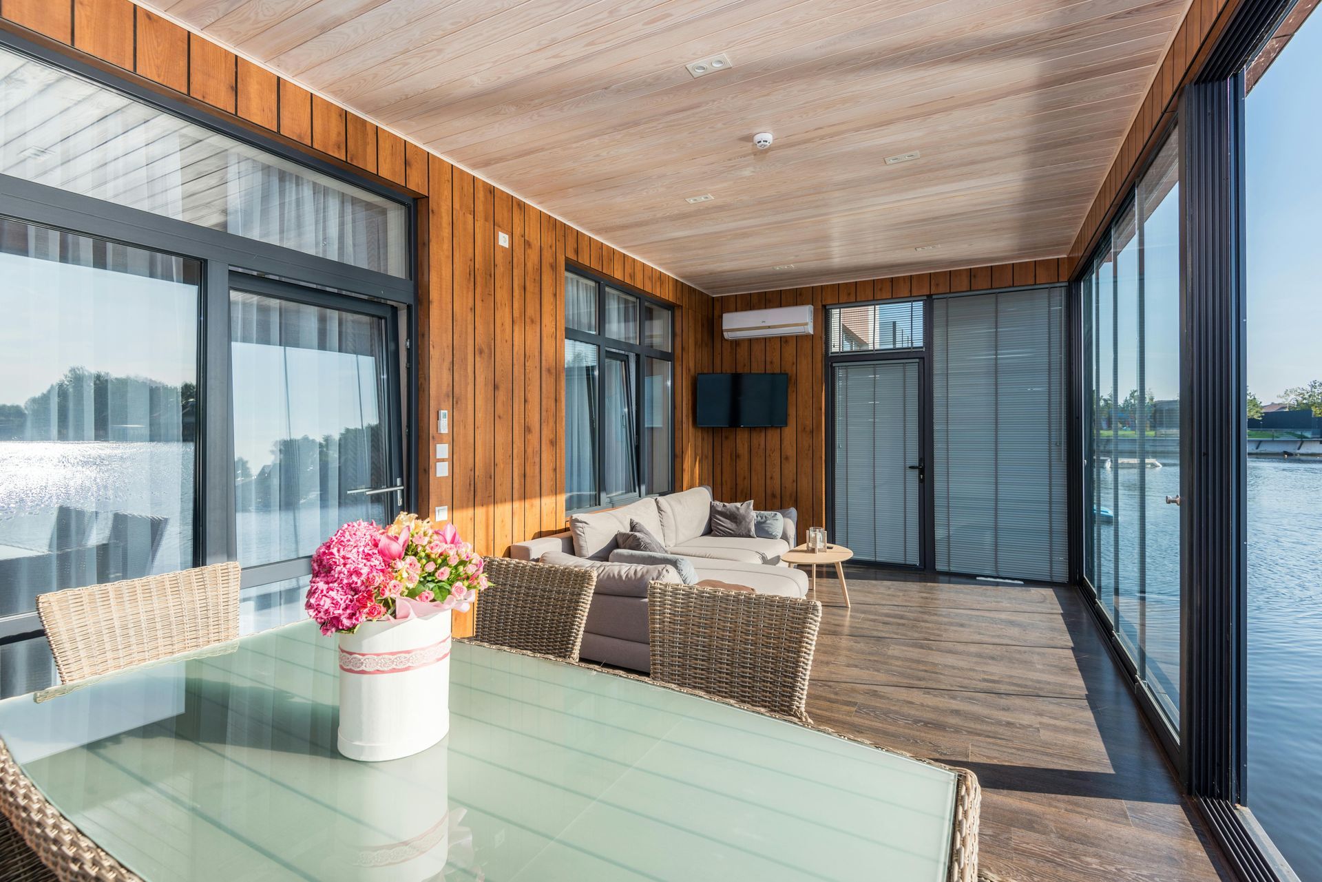 Patio with glass walls, sofa, table with flowers, wooden siding, and waterfront view.