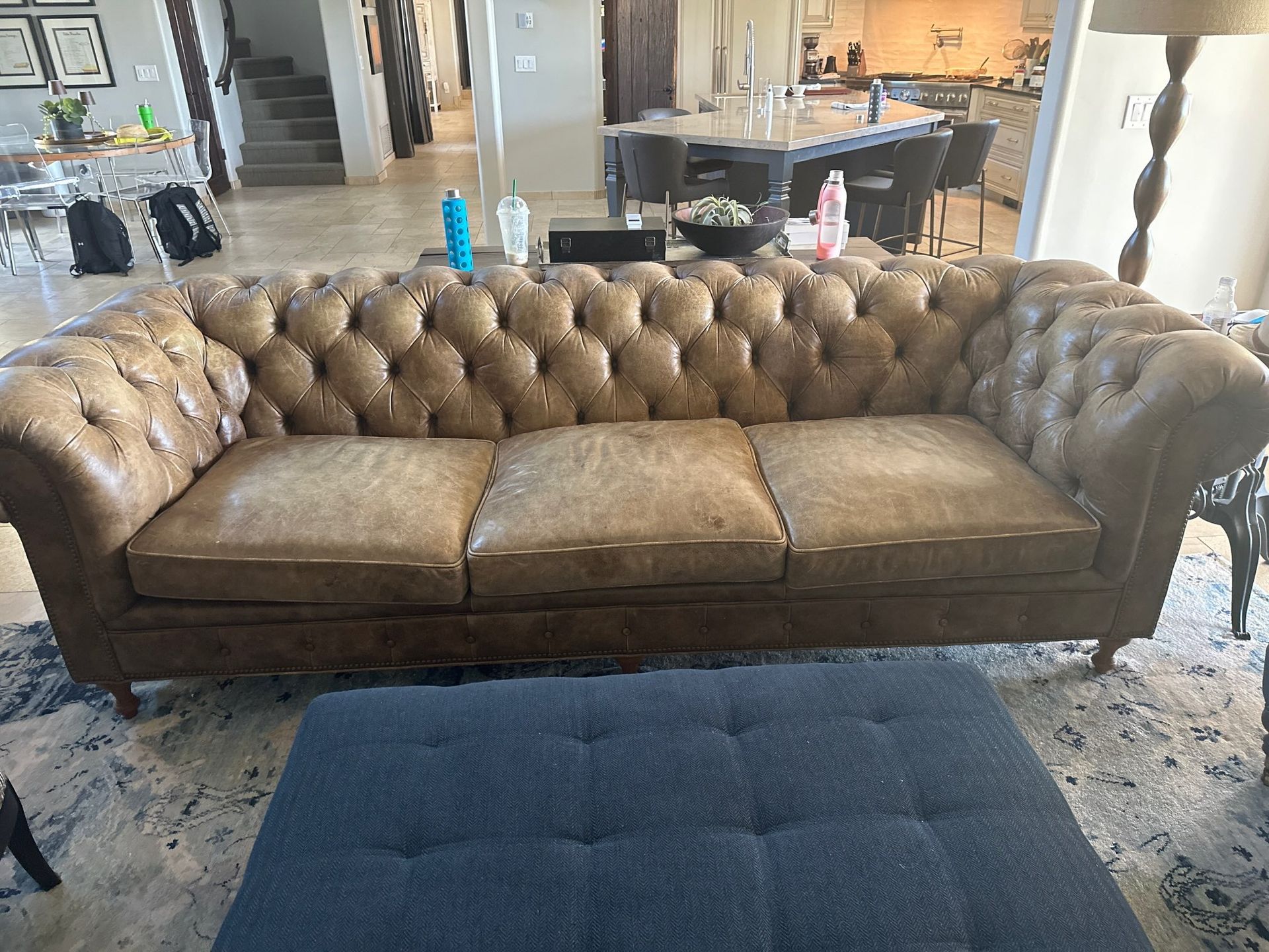 A brown leather couch is sitting in a living room next to a blue ottoman.