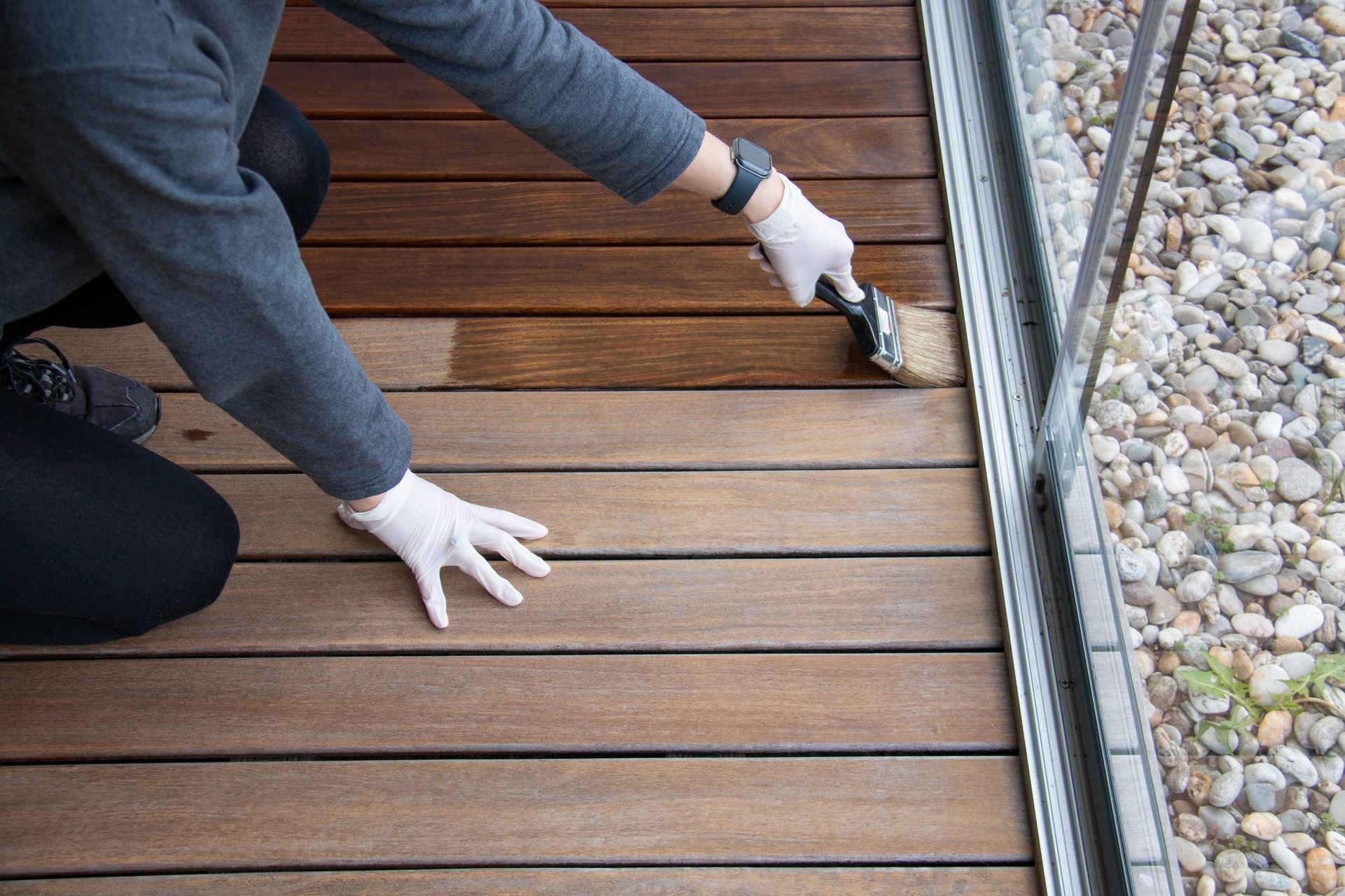 A person applying wood stain or varnish to a wooden deck using a paintbrush A person applying wood stain or varnish to a wooden deck using a paintbrush