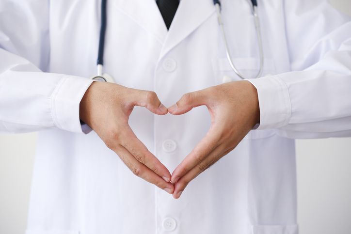 Doctor in white coat forms heart with hands, stethoscope visible, symbolizing healthcare compassion.