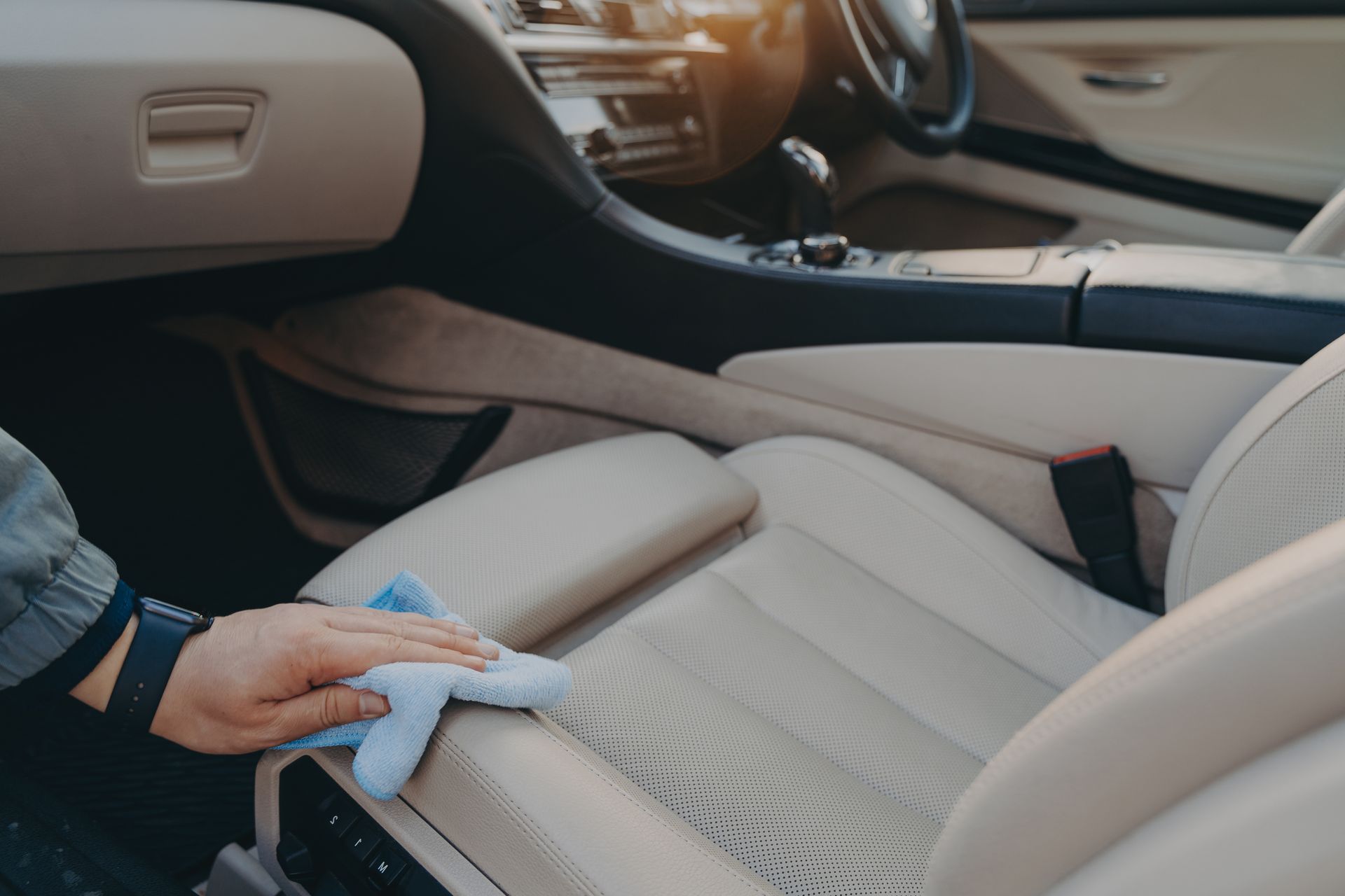 Hand cleaning beige car upholstery with a microfiber cloth inside the vehicle interior.
