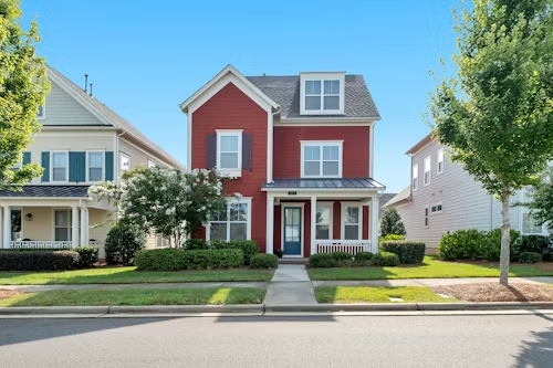 Red two-story house with a porch, flanked by green trees and adjacent homes on a sunny day.