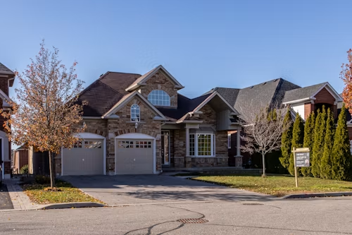 Suburban stone house with three garages and a tree-lined front yard on a sunny day