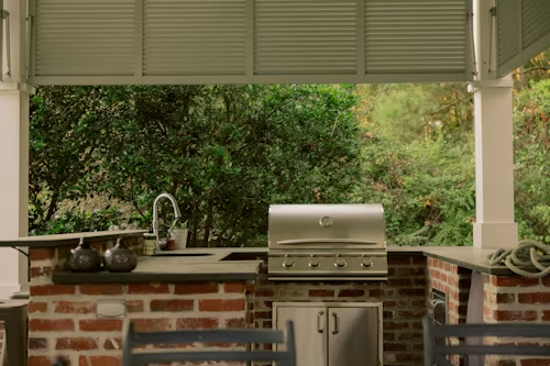 Brick outdoor kitchen with stainless grill, sink, and chairs under a covered patio overlooking greenery