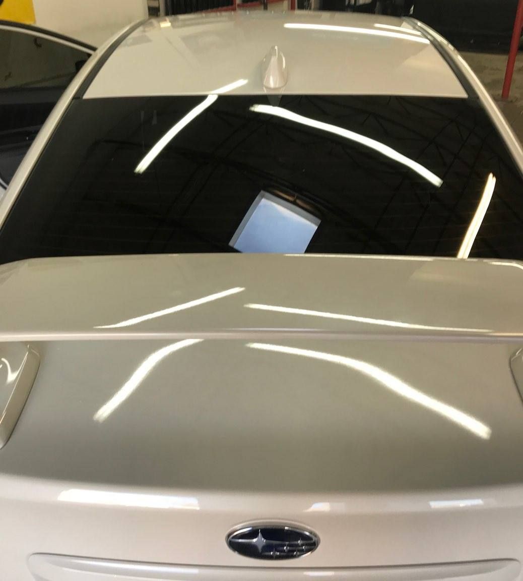 White car's rear with tinted window. Reflections on the surface and a Subaru emblem.
