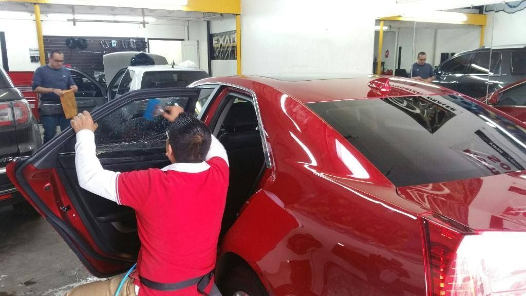 Workers tinting car windows in a shop; red car is in focus, one worker uses a tool, others watch.