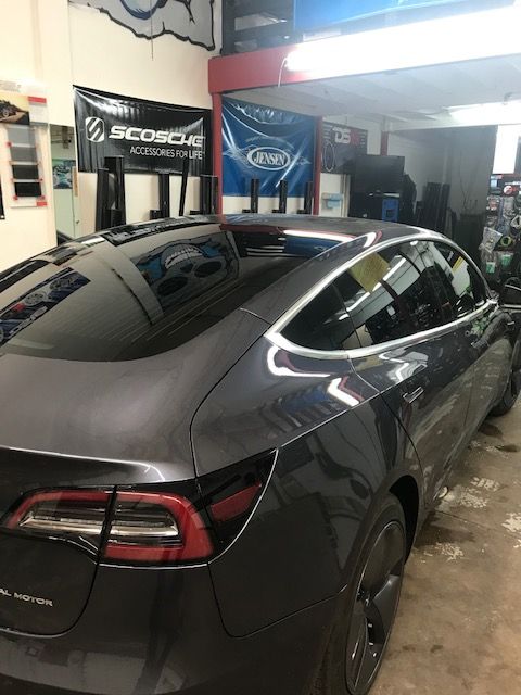Dark gray Tesla Model 3 inside a shop. Rear view with tinted windows and black rims. Two banners in the background.