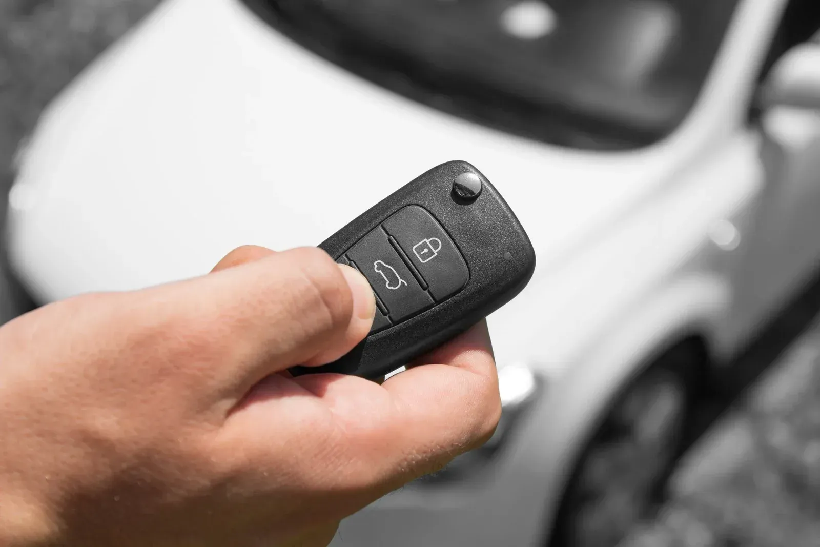 Hand holding a black car key fob, pressing the unlock button next to a white car.
