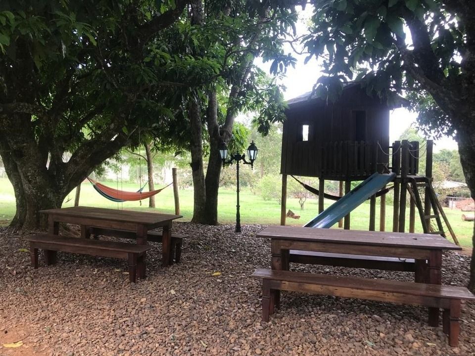 Mesas de piquenique sob árvores perto de uma casinha de madeira com escorregador.