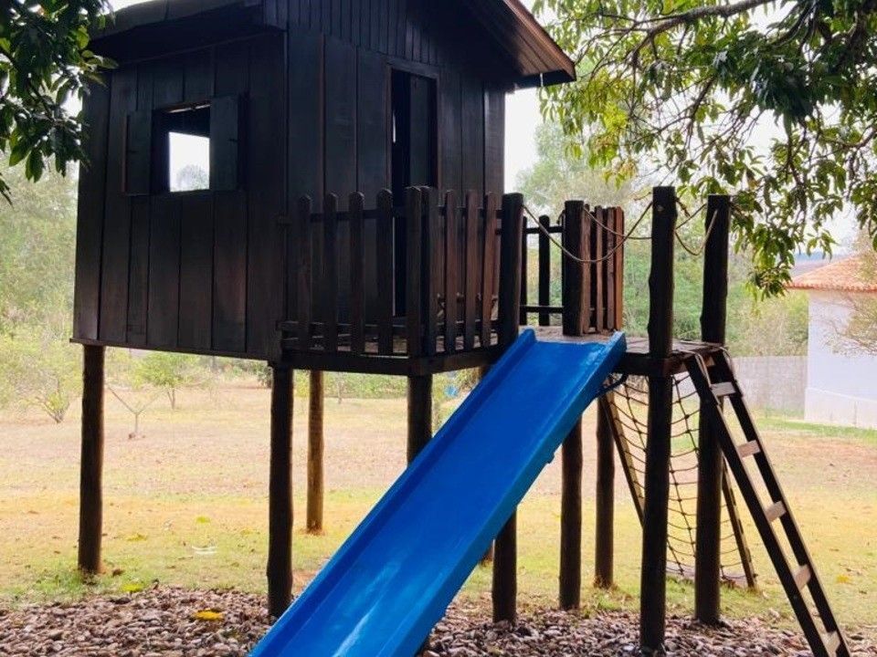 Casinha de madeira com escorregador azul, rede e escada em um quintal gramado.
