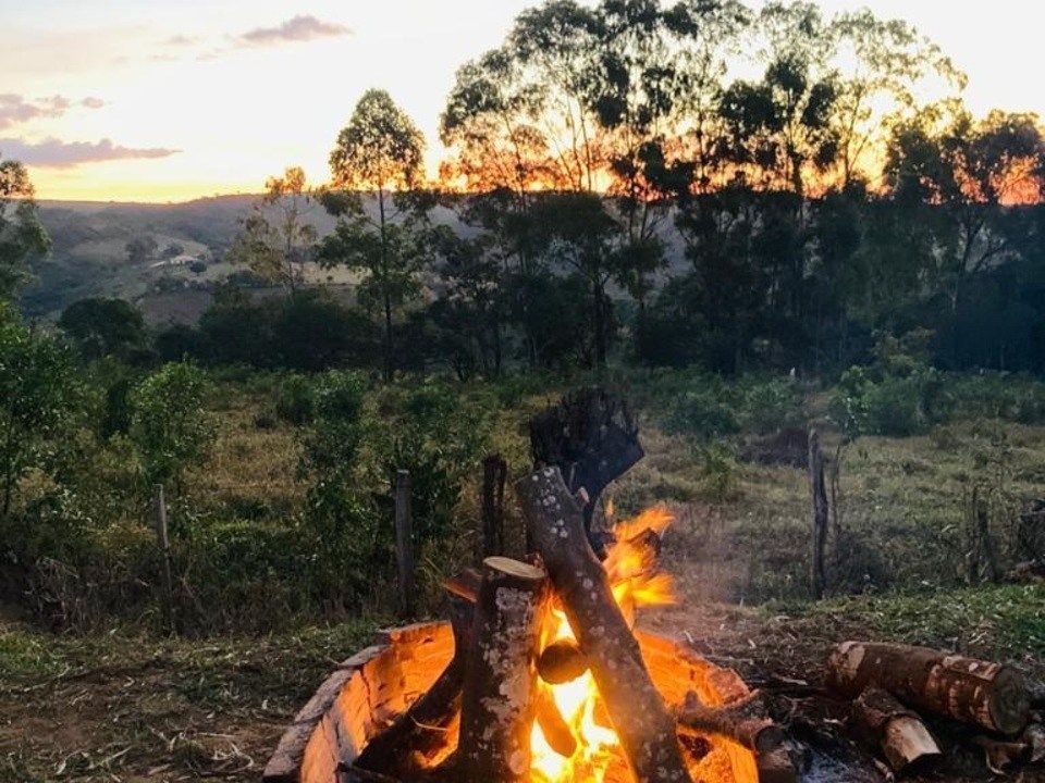 Fogueira acesa em frente a uma paisagem montanhosa ao pôr do sol, com tons de laranja e amarelo e árvores ao fundo.