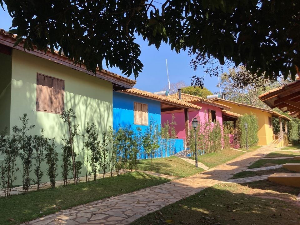 Fileira de bangalôs coloridos, cada um de uma cor vibrante diferente, com detalhes e janelas em marrom, um caminho de pedra e arbustos verdes.