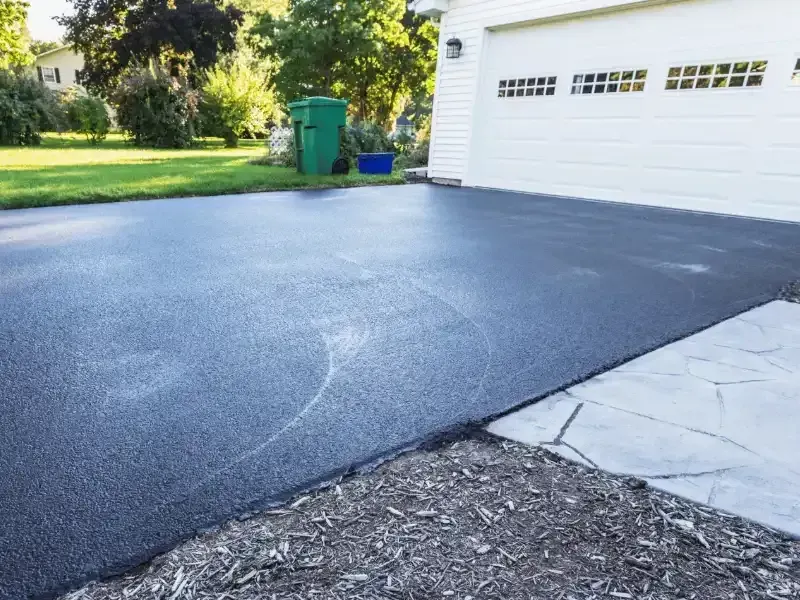 Newly sealed black asphalt driveway next to a white garage and green grass.