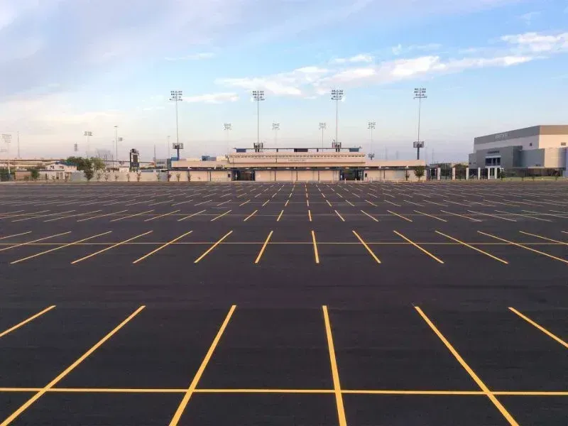 Empty asphalt parking lot in front of a stadium with bright lights under a cloudy sky.